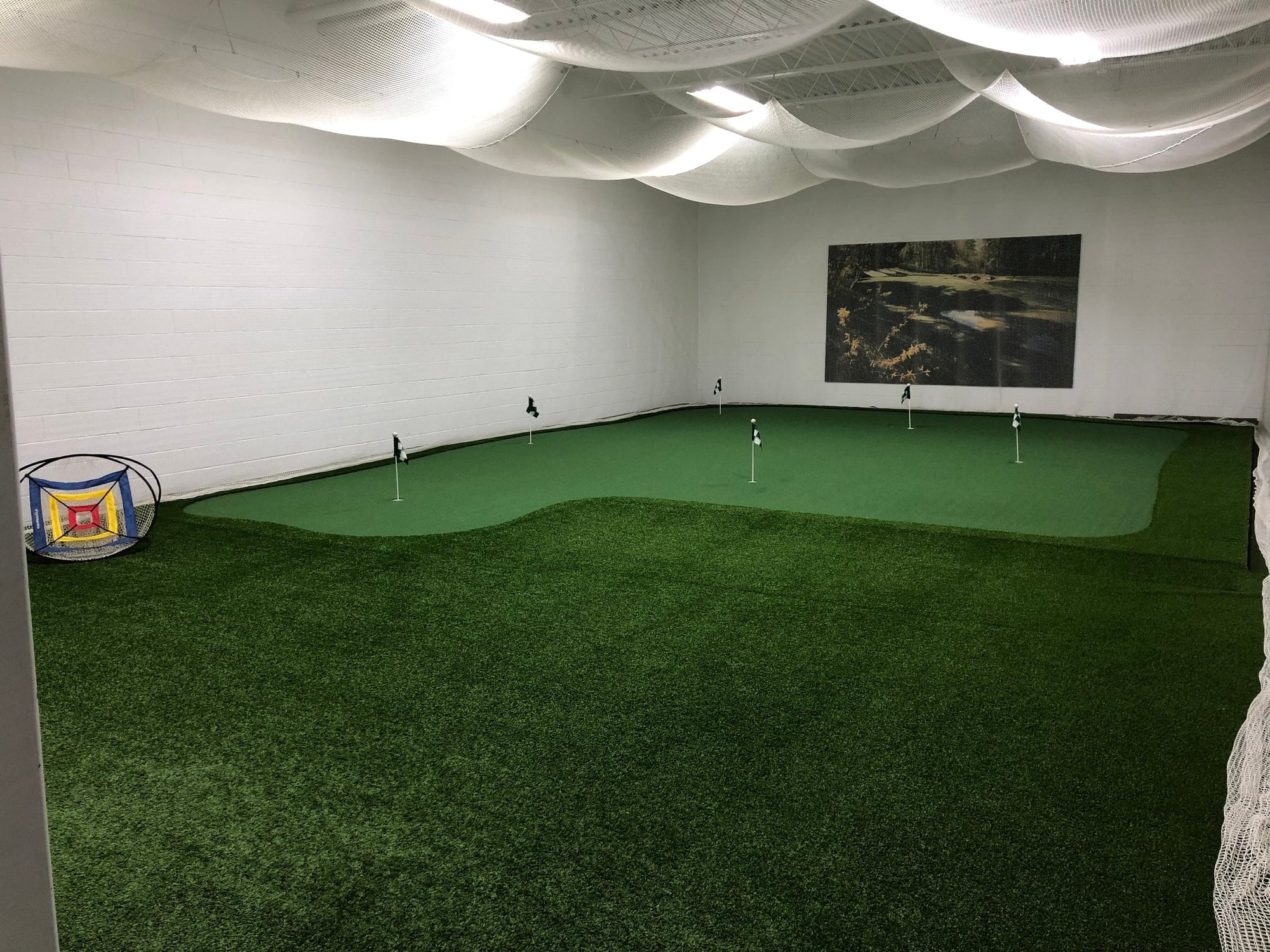 Indoor putting green with several flags and a chipping net under white ceiling netting