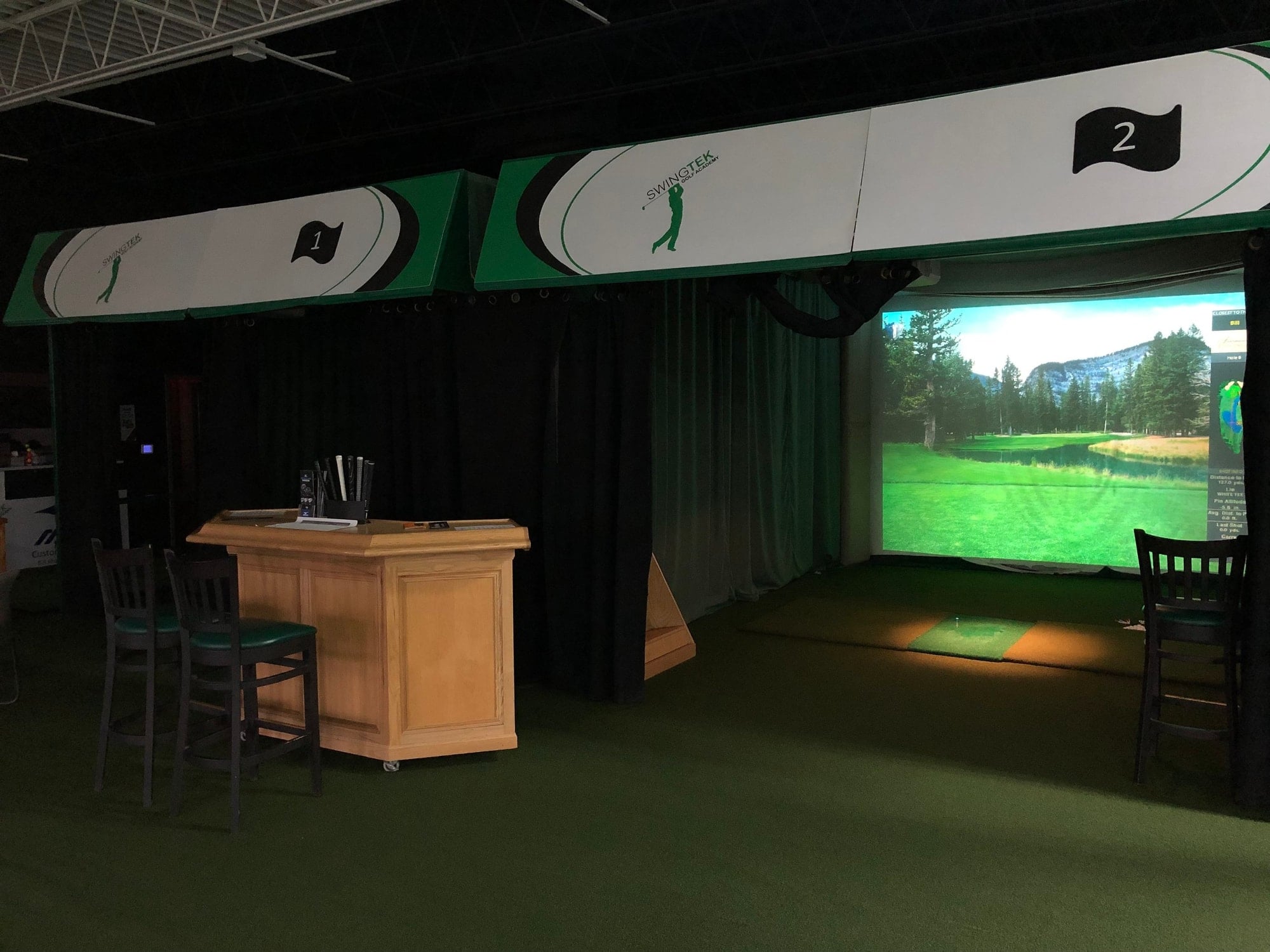 A golf simulator bay with a course on screen, a hitting mat, a wooden counter and bar chairs